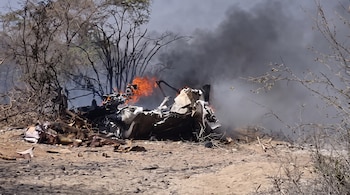 Restos carbonizados de una aeronave en llamas y humo negro se elevan desde un terreno seco con vegetación escasa y árboles