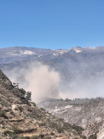 Vista panorámica de un paisaje montañoso con picos nevados, una gran nube de polvo en el valle central y cerros áridos con vegetación dispersa