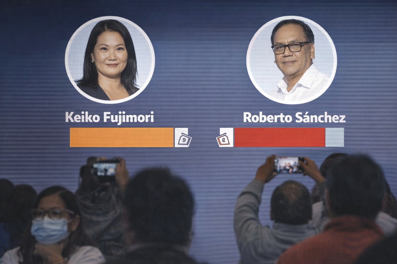 Una pantalla muestra los resultados del conteo de votos de las elecciones presidenciales, donde Keiko Fujimori aventaja ligeramente a Roberto Sánchez, mientras la audiencia captura el momento con sus teléfonos. (Imagen Ilustrativa Infobae)