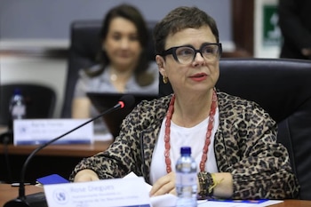 Mujer con pelo corto y gafas, chaqueta de leopardo, sentada en mesa con micrófono, botella de agua, documentos y tarjeta con nombre. Otra persona en segundo plano
