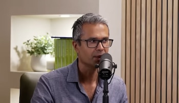 Un hombre con cabello gris y anteojos negros habla frente a un micrófono. El fondo incluye una estantería con una planta y libros, y una pared de madera