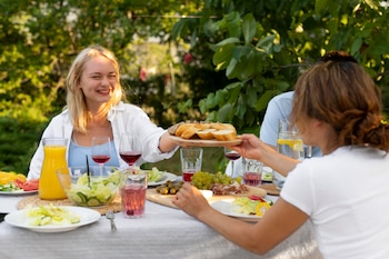 Una mujer rubia sonríe al pasar un plato de pan a otra persona en una mesa de jardín con ensaladas, frutas, zumo y vino. El fondo es de árboles