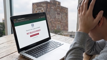 Laptop abierta en una mesa de madera. En la pantalla, resultados de examen de la UNAM con mensaje "NO SELECCIONADO". Una persona con mano en la cabeza, edificio al fondo.