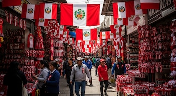 Calle concurrida de personas y tiendas con banderas de Perú, escarapelas, estandartes y decoraciones rojas y blancas.