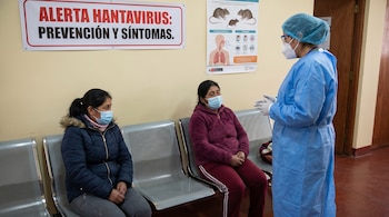 Una profesional de la salud, vestida con un traje azul y mascarilla, conversa con dos mujeres sentadas en una sala de espera. Un cartel de "ALERTA HANTAVIRUS" está detrás.