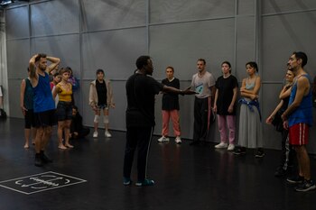 Un grupo diverso de aproximadamente quince personas, vestidas con ropa deportiva, en un amplio estudio con paredes grises y suelo oscuro, escuchando a un instructor que les habla