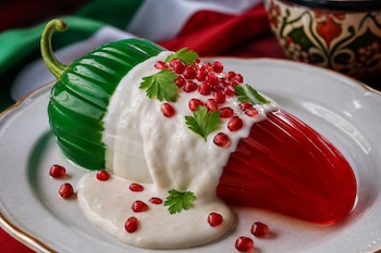 Una gelatina de colores verde, blanco y rojo con forma de chile, bañada en salsa blanca con granos de granada y hojas de perejil sobre un plato.