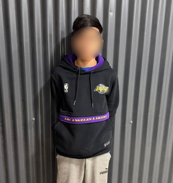 Una persona joven, de pie, con una sudadera negra de los Los Angeles Lakers y pantalones grises, contra una pared de metal corrugado gris
