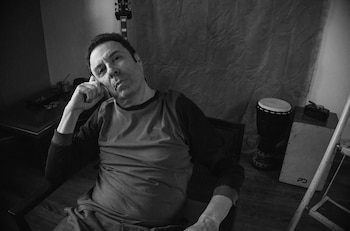 Fotografía monocromática de Mick Moss sentado, mano izquierda en la mejilla, mirando a cámara; detrás se ven una guitarra, un djembe y un cajón