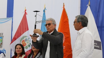 Un hombre con gafas sostiene la maqueta de una torre de monitoreo con panel solar frente a varias banderas y otras personas