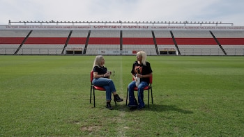 Dos mujeres sentadas en sillas rojas sobre un campo de césped. Detrás, las gradas de un estadio con asientos rojos y blancos, y nombres en la parte superior