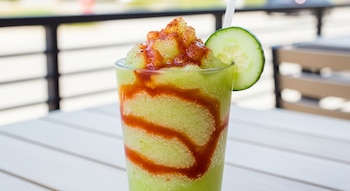 Primer plano de un frappé verde con jarabe de chamoy rojo en espiral y chile en polvo, decorado con una rodaja de pepino y una pajita blanca.
