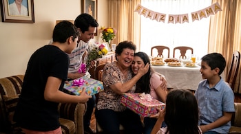 Madre colombiana sonriente sentada en un sofá, abrazando a una joven y recibiendo regalos y flores de su familia en una celebración del Día de la Madre.