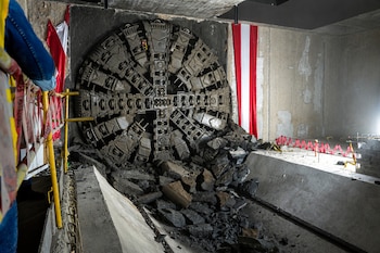 Gran cabeza de tuneladora gris emerge de una pared de tierra en una estación subterránea, con rocas excavadas. A los lados, banderas peruanas y barreras de seguridad