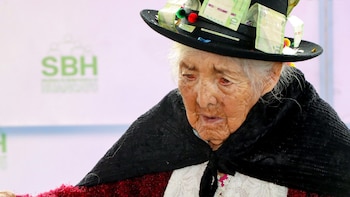 Primer plano de una mujer anciana con piel arrugada y pelo blanco, con un sombrero negro decorado con billetes de S/10 y pompones de colores
