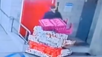 Una figura borrosa con ropa oscura frente a una pared roja y un objeto voluminoso que parece ser un medidor o equipo electrónico