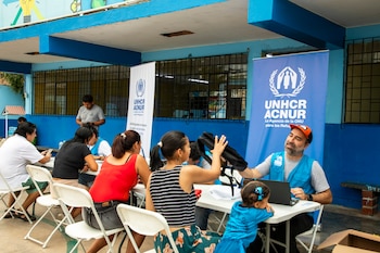 Personas sentadas en mesas de una cafetería, interactuando con un representante de ACNUR que usa una laptop, con banderas de la organización al fondo
