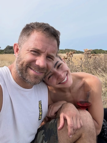 Thelma Fardin sonríe junto a un hombre barbudo, ella recostada en su hombro. Él lleva camiseta blanca con logo Edikted. Ella sostiene copa de vino. Fondo de campo y cielo
