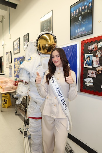 Una mujer con traje claro y banda de Miss Universe posa con los pulgares arriba al lado de un traje de astronauta con casco dorado