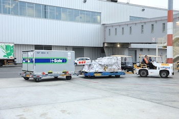 Vehículo remolcador con conductor arrastra plataforma con contenedor blanco y otra con bultos embalados en aeropuerto