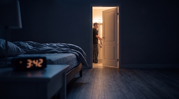 Un dormitorio a oscuras con un reloj digital que marca las 3:42 y un hombre con pantalón de pijama frente al baño iluminado.