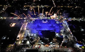 Vista aérea nocturna de una plaza llena de multitud iluminada en azul, rodeada de calles de la ciudad y fuegos artificiales en el cielo