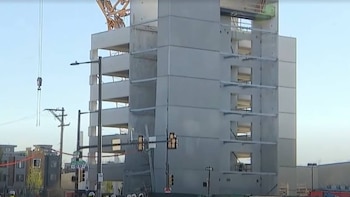 Vista exterior de un edificio de gran altura en construcción con múltiples pisos y ventanas abiertas, una grúa de construcción y señales de tráfico