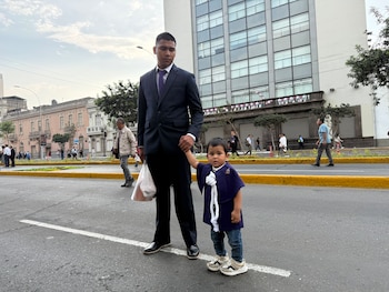 Un hombre de traje oscuro toma la mano de un niño con túnica morada y cordón blanco en medio de una avenida con edificios al fondo
