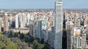 Vista aérea de una ciudad con edificios de diversas alturas, un parque arbolado con vehículos estacionados y un gran mural de Lionel Messi en la fachada de un edificio