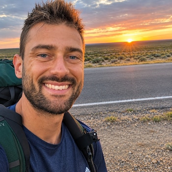 Primer plano de un hombre sonriente con barba y mochila junto a una carretera asfaltada frente a una puesta de sol en un terreno llano