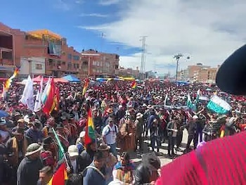 Vista aérea de una gran multitud de personas, algunas ondeando banderas coloridas, reunidas en una plaza al aire libre con edificios y un cielo azul de fondo