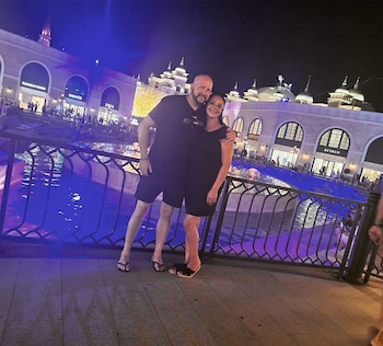 Un hombre y una mujer sonriendo posan de noche frente a una barandilla metálica, un canal de agua azul y edificios comerciales con arcos y luces