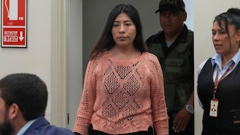 Mujer de cabello oscuro con suéter calado salmón, un policía con chaleco, otra mujer con chaleco oscuro y un cartel de extintor en una pared blanca
