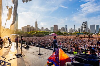 Escenario de concierto al aire libre con banda tocando, multitud de personas, torres de altavoces y luces, y un paisaje urbano con rascacielos bajo un cielo con nubes