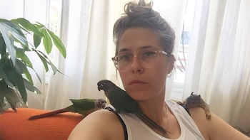 Natasha Newman-Thomas, mujer con gafas y cabello recogido, tiene dos loros y una ardilla en sus hombros. Al fondo, cortinas claras y una planta
