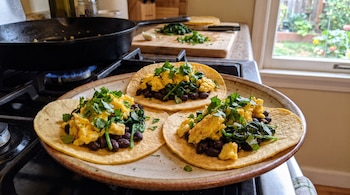Tres tortillas con frijoles negros, huevo revuelto, espinaca y cilantro en un plato sobre una estufa con una sartén al fondo.