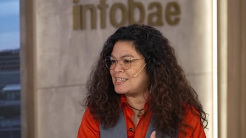 Primer plano de una mujer con cabello rizado oscuro, gafas y un micrófono, vestida con camisa naranja y chaleco gris, frente al logo de Infobae