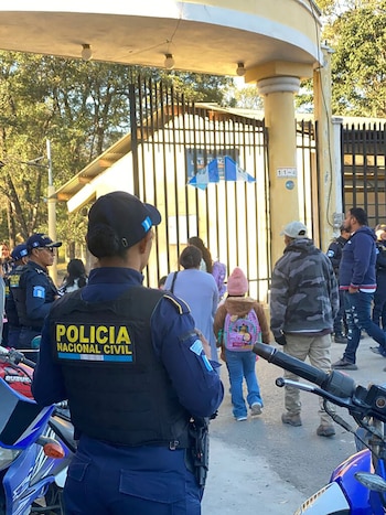 Vista trasera de una agente de la PNC de Guatemala con uniforme azul y chaleco, supervisando la entrada de una escuela. Hay otras personas y niños en el fondo