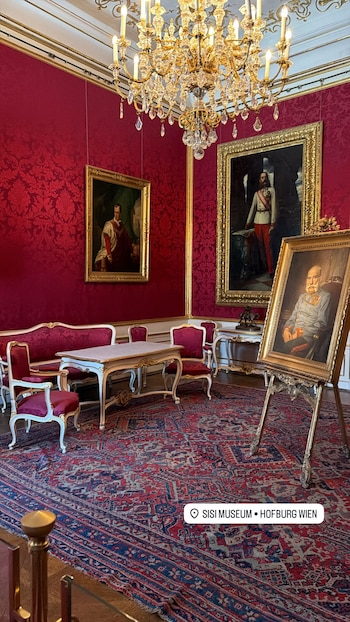 Interior de un salón en el Museo Sisi de Viena con paredes rojas damascas, cuadros históricos, muebles dorados y tapizado rojo, y una lámpara de araña