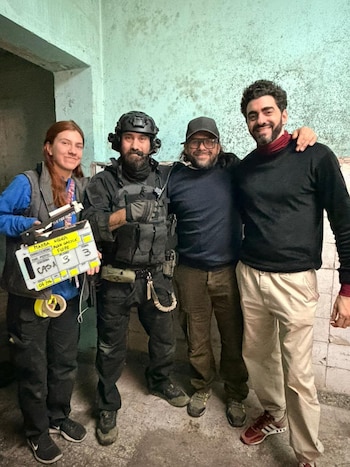 Cuatro personas sonrientes posan en un interior con paredes de color cian. Una mujer sostiene una claqueta con "MAMBA NEGRA". Un hombre lleva equipo táctico