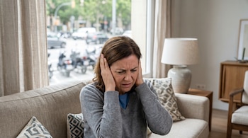 Mujer de mediana edad sentada en un sofá claro, cubriéndose los oídos con ambas manos, con expresión de estrés. Detrás, una ventana muestra tráfico urbano borroso.