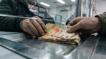 Manos sujetan billetes de mil y quinientos pesos argentinos sobre una bandeja metálica ante un cristal.