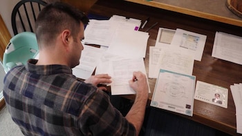 Un hombre de espaldas, vestido con camisa a cuadros, escribe en un formulario. Sobre la mesa de madera hay muchos documentos, certificados y una foto de pasaporte