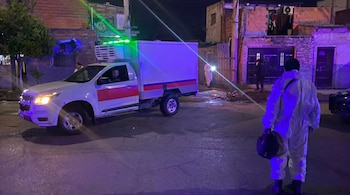 Un camión blanco refrigerado estacionado de noche en una calle urbana con edificios de ladrillo. Varias personas, una con traje protector, están presentes.