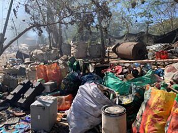 Vista cercana de una gran acumulación de residuos, incluyendo bolsas de basura de colores, escombros y un objeto metálico oxidado en un entorno abierto con árboles y humo