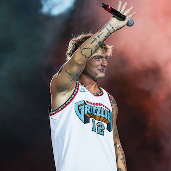 Un hombre rubio con tatuajes en los brazos lleva una camiseta blanca sin mangas y levanta un micrófono negro. El fondo es oscuro con humo rojizo