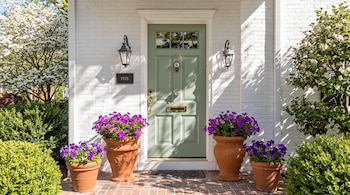 Puerta verde en una pared de ladrillo blanco con cuatro macetas de terracota con flores moradas, dos faroles y arbustos.