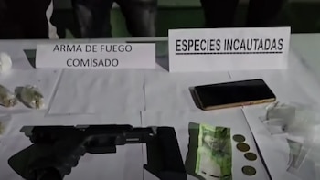 Una mesa blanca muestra una pistola, un celular, billetes, monedas, varias bolsas pequeñas con sustancias y dos carteles: 'Arma de fuego comisado' y 'Especies incautadas'
