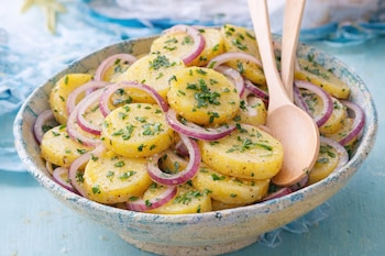 Un cuenco con ensalada de patata y cebolla roja, decorada con hierbas verdes picadas, y dos cucharas de madera.