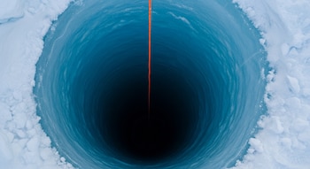 Fotografía aérea de un pozo profundo en hielo azul. Una cuerda naranja desciende verticalmente hacia el interior oscuro, rodeado por nieve texturizada.
